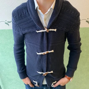 Mörkblå kofta från L.O.G.G. med snörning - Mysig mörkblå stickad kofta från L.O.G.G. med snygga träknappar och vit snörning framtill. Koftan har ett coolt mönster upptill, bred krage och långa ärmar. Perfekt för lager-på-lager och chill stil.