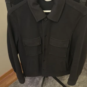 Svart overshirt från Zara med fickor - Svart overshirt från Zara i storlek S. Jackan har klassisk krage, två stora bröstfickor med lock och knäppning framtill. Perfekt för lager-på-lager och enkel att styla till olika outfits.