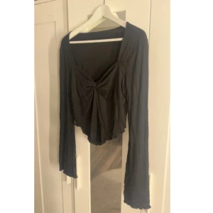 Svart långärmad croppad topp - Svart långärmad topp med croppad passform och twistad knutdetalj framtill. Toppen har v-ringning och vida ärmar som ger en cool och avslappnad vibe. Perfekt att styla med höga jeans eller kjol.