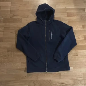 Marinblå hoodie från Sail Racing - Marinblå hoodie från Sail Racing med hel dragkedja och huva. Tröjan har en bröstficka med dragkedja och Sail Racing-logga. Tillverkad i 65% bomull och 35% polyester för en skön och slitstark känsla. Perfekt för kyliga dagar.