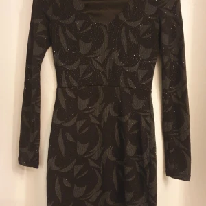 Svart glittrig kortklänning från Voyelles - Säljer en svart kortklänning med glittrigt abstrakt mönster. Klänningen har långärmade ärmar och en snygg v-ringad hals.
