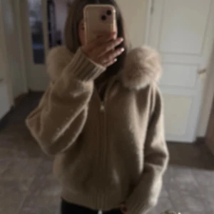 Beige hoodie med päls från Alessa - Mysig beige hoodie från Alessa med fluffig päls runt huvan. Tröjan är stickad, har dragkedja framtill och långa ärmar med breda ribbade muddar. Perfekt för kalla dagar när du vill vara både varm och snygg.