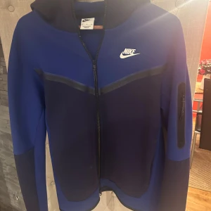 Blå Nike hoodie med dragkedja S - Nike hoodie i två nyanser av blått med hel dragkedja och huva. Snygg sportig look med svart detalj över bröstet och praktisk ficka med dragkedja på ärmen. Perfekt för dig som gillar streetwear och sportig stil.