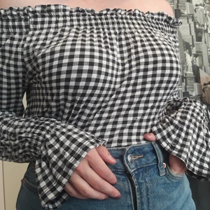 Rutig topp - Svart och vit rutig off-shoulder tröja. Små volanger längs "kragen" och handlederna, har även resårband på samma ställen. Storlek: S/M. Material: Viskos. Väldigt lätt och luftig topp. Två små hål, ett på handvolangen och ett på framsidan annars inga skador.