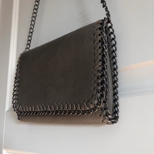 Väska med kedja - Helt onanvänd grå fejkskinnsväska med snygg kedjedetalj. Höjd 15cm bredd 22cm. Går att korsa banden inuti som jag gjort på sista bilden om man vill ha de kortare. Inköpt för 300kr säljer då det inte riktigt är min stil. Frakt 66kr    BUD:80kr
