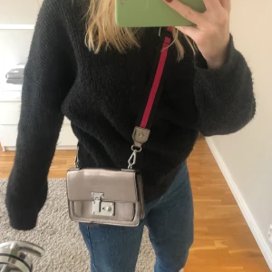Väska - Jätte cool silvrig väska med rosa/svart tillhörande band. Bandet går att ta av. Väskan är i mycket bra skick, använd ett fåtal gånger. Har justerbart band så man kan göra den både kortare och längre.                                                                                  Köparen står för frakt.
