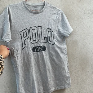 Grå Ralph Lauren T-shirt  - En grå Polo Ralph Lauren T-Shit med mörkblått tryck på magen. Tröjan är lite tjockare och mycket skön. Passar både tjej och kille. 