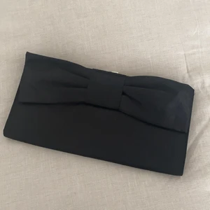 Kuvertväska  - Söt, stel kuvertväska med svart rosett i lätt glansigt material. Använd två kvällar. I väskan finns hållare för kedja som du kan koppla på om du har en eller köper till. 