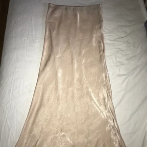 Beige lång kjol från boohoo, strl 36 - Beige lång kjol i satin liknande material. Kjolen kommer från boohoo och är i storlek 36. Kjolen är nästan aldrig använd och är i väldigt bra skick, den är dock aningen genomskinlig.