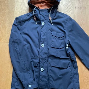 Vår-  och höstparkas  - En jacka i parkasmodell i storlek M från märket Elvine. Marinblå med vita knappar och andra detaljer. Foder i nät  i en brunorange färg. Är i mycket bra skick. Inga skador eller missfärgningar. Använd ett fåtal gånger. Är inte tvättad. Nypris ca 1600 kr. 