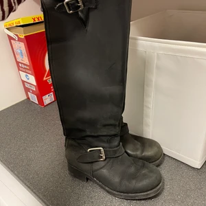 Stövlar höga svarta  - Stövlar som går till knäna. Svarta i skinn med spännen uppe och nere, relativt gott skick. Dragkedjor baktill. Kan bli i ännu bättre skick om man smörjer dem!