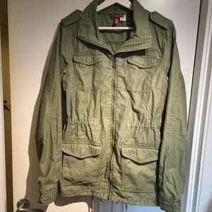 Grön cargojacka från H&M - Grön bomullsjacka från H&M. Fått av en kompis men aldrig använt själv. Den har en fläck på ena fickan, se bild 3. Är i storlek 42 men skulle säga att den passar som en M. Den har snörning vid midjan samt längst ner, så man kan göra den smalare. 
