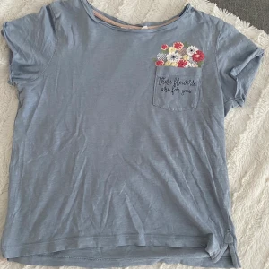 Blå t-shirt med blommig ficka  - En vanlig ljusblå t-shirt, med en blommig ficka. Köpare står för frakt!