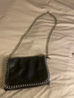 Svart axelväska med kedjerem - Snygg svart axelväska i fejkläder med grov silverfärgad kedja som axelrem och detaljer runt kanterna. Väskan har magnetlås och innerfack med dragkedja. Perfekt för dig som gillar en edgy och trendig stil.