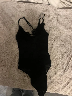 Svart spetsbody med tunna axelband - Säljer en svart body i spets med tunna justerbara axelband och v-ringning. Bodyn har ett blommigt spetsmönster och fina spetsdetaljer vid urringningen. Aldrig använt, stretch material. Snygg att ha under jeans, banden på rygggen går även i kors!