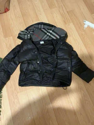 Svart pufferjacka med rutig huva - Säljer en svart pufferjacka med kortare passform och stor huva fodrad med klassiskt rutigt mönster. Jackan har dragkedja och knappar framtill samt två sidofickor. Perfekt för kalla vinterdagar och riktigt snygg streetstil.