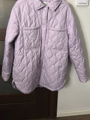 Ljuslila quiltad pufferjacka - Säljer en ljuslila pufferjacka med quiltat mönster och två stora bröstfickor. Jackan har krage, tryckknappar framtill och en oversized passform. Perfekt för kalla dagar och ger en trendig look med sin pastellfärg och vadderade design.
