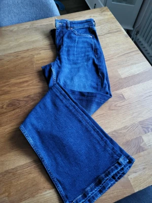 Blå bootcut jeans med utsvängda ben - Säljer ett par klassiska blå bootcut jeans med utsvängda ben och normal midja. Jeansen har fem fickor och stängs med dragkedja och knapp. Perfekta för dig som gillar retrostil och vill ha ett par bekväma jeans i tidlös modell.