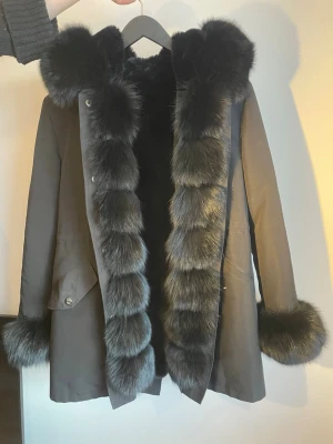Svart parka jacka med äkta päls  - Snygg svart pälsjacka med fluffig äktapäls längs huva, ärmslut och hela framsidan. Jackan har en rak passform, men går att knyta runt midjan. stora fickor och stängs med knappar. Perfekt för kalla vinterdagar när du vill hålla dig varm och se trendig ut. Pris kan diskuteras