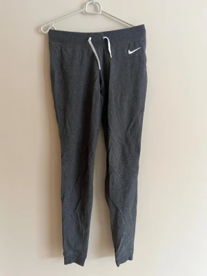 Mörkgrå mjukisbyxor från Nike - Snygga mörkgrå mjukisbyxor från Nike med vit dragsko i midjan och klassisk Nike-logga på vänster sida. Byxorna har smal passform och muddar vid bensluten. Perfekta för chill eller träning, tillverkade i mjukt och stretchigt material.