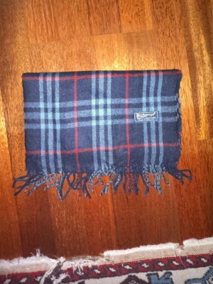 Mörkblå rutig halsduk från Burberry - Klassisk halsduk från Burberry i mörkblått med rutigt mönster i rött och grått. Halsduken har fransar i kanterna och en vävd Burberry-etikett. Perfekt accessoar för att lyfta din outfit med en tidlös look.