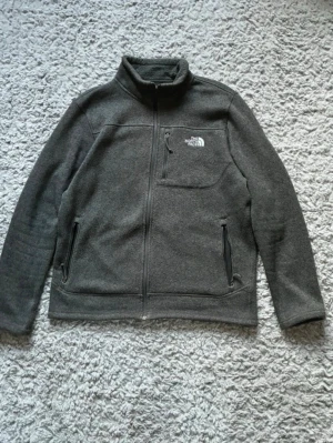 Grå fleecejacka från The North Face - Mysig grå fleecejacka från The North Face med dragkedja framtill, hög krage och tre fickor med dragkedja. Perfekt för lager-på-lager och håller dig varm under kyliga dagar. Klassisk logga på bröstet och enkel, stilren design.