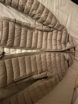 Beige dunjacka med dragkedja - Lätt och stilren beige dunjacka med horisontella quiltade partier, två fickor med dragkedja och hög krage. Jackan har hel dragkedja framtill och är perfekt för kalla vinterdagar. Den har bara använts ett fåtal gånger så den är väldigt ny.