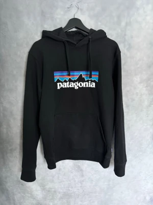 Svart Patagonia hoodie med tryck - Snygg svart hoodie från Patagonia med det klassiska färgglada logotrycket på bröstet. Riktigt bra skick då den knappt är använd.