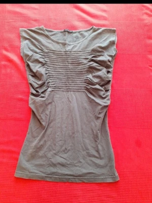 Grå topp med rynkade detaljer - Snygg grå topp med rynkade detaljer och horisontella sömmar över bröstet. Modellen är ärmlös och har en rund halsringning. Perfekt till jeans eller kjol för en clean och modern look.