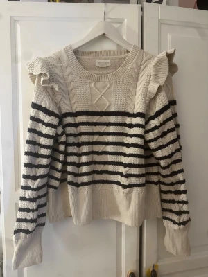 Randig stickad tröja med volang - Beige stickad tröja från Holly & Whyte by Lindex med svarta horisontella ränder, volangdetaljer på axlarna och kabelstickat mönster framtill. Tröjan har rund halsringning och långa ärmar. Perfekt för dig som gillar en söt och trendig look.