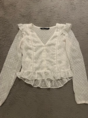 Vit prickig blus med volang från Zara Stockholmsstil  - Supersöt vit blus från Zara med små prickar, volangkant nertill och dekorativa sömmar framtill. Blusen har v-ringning, långa ärmar i transparent tyg och fina volangdetaljer vid axlarna. Perfekt för en trendig och romantisk look. Den är inte kritvit utan mer benvit