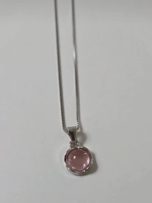 Silverhalsband med rosa sten - Snyggt halsband i silverfärg med en tunn kedja och ett runt hänge med en ljusrosa, glansig sten infattad i metall. Stenen har en mjuk, pärlliknande yta som ger ett elegant intryck. Perfekt accessoar för att lyfta din outfit. 45 cm + 5 cm förlängningskedja.