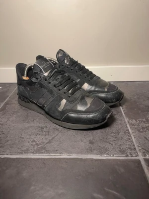 Svarta camo sneakers från Valentino - Snygga svarta sneakers från Valentino med diskret camo-mönster och svarta skosnören. Skorna har en låg siluett och är tillverkade i en mix av läder och textil. Perfekta för dig som vill ha en stilren men ändå unik look.