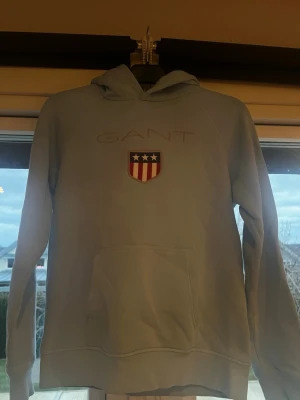 Ljusblå hoodie från GANT - Snygg ljusblå hoodie från GANT med broderad logga och ett sköldmärke med stjärnor och ränder på bröstet. Hoodien har en stor känguruficka framtill och klassisk huva. Perfekt för en avslappnad och sportig stil.