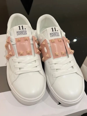 Valentino Rockstud sneakers vit/rosa - Snygga vita Valentino Rockstud sneakers med rosa detaljer och ikoniska nitar. Skorna har rund tå, platt sula och snörning. Tillverkade i skinn med rosa läderpaneler och matchande nitar för en edgy look. Perfekta för dig som vill sticka ut.