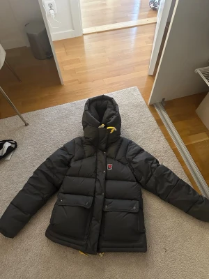 Svart dunjacka från Fjällräven - Snygg svart dunjacka från Fjällräven med stor huva och gula detaljer på dragkedjan. Jackan har två stora framfickor med lock och tryckknappar samt Fjällrävens klassiska rävlogga på bröstet. Perfekt för kalla dagar och riktigt varm.