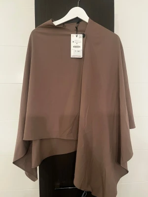 Zara Brun cape i storlek M - Säljer en brun cape-kappa från Zara. Kappan har en asymmetrisk design med knäppning vid halsen och faller snyggt över axlarna. Tillverkad i ett tunt, mjukt tyg som ger ett elegant och modernt intryck.