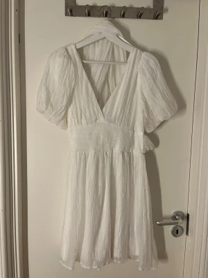 Vit studentklänning - Supersöt vit studentklänning med puffärmar. Klänningen har v-ringning, markerad midja och knytband i ryggen för extra detalj. Tillverkad i ett lätt, strukturerat tyg som ger en luftig känsla. Perfekt för sommarens alla tillfällen!