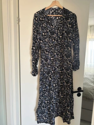 Blommig långklänning från Ellos - Snygg långklänning från Ellos med svart botten och blommigt mönster i vitt och ljusblått. Klänningen har v-ringning och långa ärmar, perfekt för dig som gillar en feminin och avslappnad stil. Tillverkad i mjuk och skön viskosblandning.