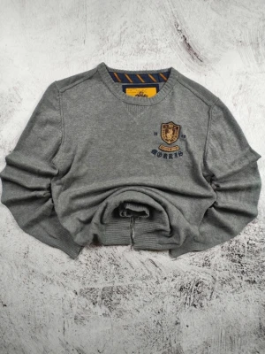 Grå tröja med emblem från Morris - Snygg grå tröja från Morris med broderat emblem och text på bröstet. Tröjan har rund halsringning och ribbade muddar. Perfekt för dig som gillar klassisk stil med en twist. Materialet känns mjukt och bekvämt.