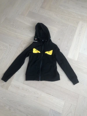 Svart hoodie med gula ögon från Fendi - Säljer en svart hoodie från Fendi med dragkedja och huva. På bröstet finns två gula ögon som ger ett coolt och unikt uttryck. Tillverkad i Italien och har en klassisk passform med ribbade muddar. Perfekt för dig som vill sticka ut i mängden.