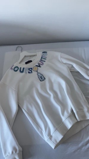 Vit  sweatshirt - Säljer en vit sweatshirt från h&m  med färgglad logotyp och tryck framtill. Tröjan har rund halsringning, långa ärmar och ribbade muddar. Materialet känns mjukt och skönt, perfekt för en clean och trendig look.