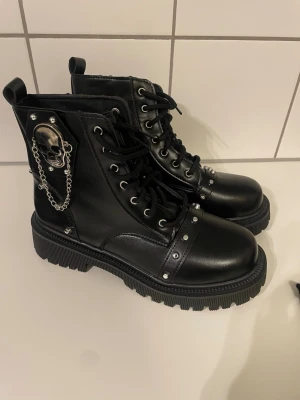 Svarta boots med dödskalle och kedja - Tuffa svarta boots i syntetläder med snörning, silverfärgade nitar och en stor metallisk dödskalle med kedja på sidan. Grov sula och högt skaft ger en edgy look. Perfekta för dig som gillar alternativ stil.