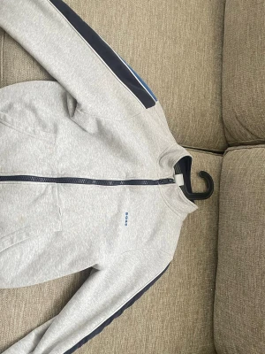 Grå ziptröja från BOSS - Snygg grå ziptröja från BOSS med marinblå detaljer längs ärmarna och diskret BOSS-logga på bröstet. Tröjan har hög krage, dragkedja framtill och praktiska fickor. Perfekt för en sportig och avslappnad stil.