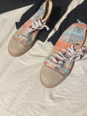 Chanel färglada skor - Unika höga sneakers från Chanel med färgglatt mönster i orange, blått och beige. Skorna har flätad tå i naturmaterial, textilovandel och beige snörning. Sula i gummi med Chanel-logga undertill och guldfärgade öljetter. Perfekta för dig som vill sticka ut.