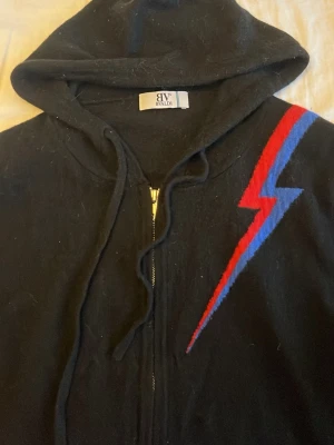 Svart hoodie med blixt från BVALDI  - Svart hoodie med dragkedja från BVALDI. På bröstet finns en cool blixt i rött och blått som ger en unik vibe. Hoodien har huva med dragsko och ribbade muddar. Passar dig som är runt 180. Kostar runt 1300kr ny och mitt pris är endast 799. Använd fåtal gånger då den inte passa just mig.
