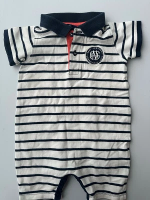 Randig kortärmad jumpsuit med krage storlek 74 - Kortärmad jumpsuit i vitt med mörkblå horisontella ränder och mörkblå krage. Knäppning framtill med två knappar och en liten röd detalj vid knappslån. Tryckt emblem på bröstet. Mjuk bomullskänsla och sportig look.