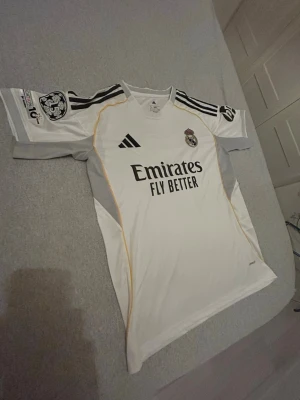 Real Madrid matchtröja Mbappé 10 - Säljer en vit Real Madrid matchtröja från Adidas med svarta detaljer och gula linjer. Tröjan har trycket 'Emirates Fly Better' på framsidan, klubbmärke och Adidas-logga. På ryggen står det 'Mbappé' och nummer 10. Korta ärmar och CL-märke på axeln.