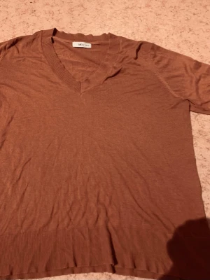 Brun v-ringad topp från Ellos - En brun v-ringad topp från Ellos med korta ärmar och ribbade detaljer vid halsringning och nederkant. Toppen har en loose passform och är tillverkad i ett mjukt material som känns skönt mot huden. Perfekt att styla med jeans eller kjol.