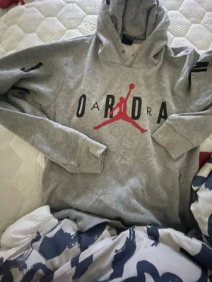 Grå Jordan hoodie med tryck - Snygg grå hoodie från Jordan med det klassiska Jumpman-loggan i rött framtill och svart text. Hoodien har huva och långa ärmar, samt ribbade muddar vid ärmslut och nederkant. Perfekt för en sportig och avslappnad stil.